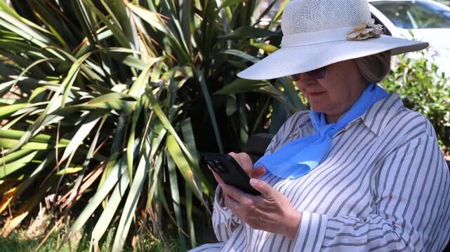 Senior Woman Using Smartphone Outdoors. Senior woman in sun hat using smartphone outdoors near tropical plants. Mature traveler checking phone during vacation or city walk.
