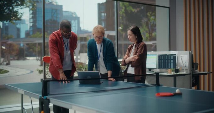 Three Software Developers Gather Around a Laptop on a Ping Pong Table for Office meeting, Coding App Features. Youthful and Professional Team of Engineers Working on Cybersecurity Solutions.