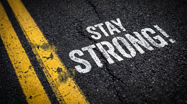 The motivational phrase "STAY STRONG!" is painted in textured white lettering on a cracked asphalt road surface right next to double solid yellow lines.