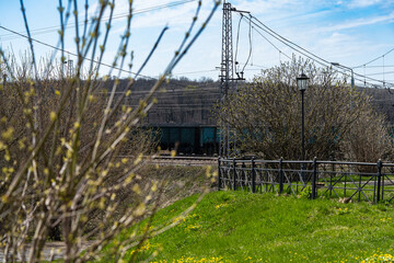 Fototapeta premium Freight train moves through the landscape, partially hidden by budding trees with a bright green foreground. Railway lines and infrastructure are visible, creating a dynamic scene.