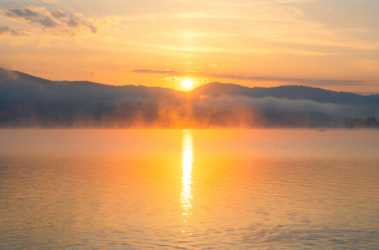 Sun rising over mountains with mist on a calm lake and a glowing golden reflection on the water