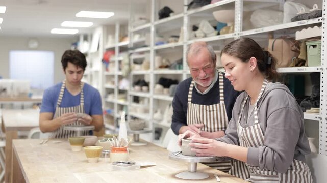 Eldery man helps and gives instructions to young woman sculpting clay at workbench in ceramic workshop