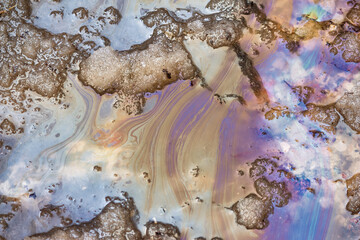 Fototapeta premium Iridescent oil slick patterns swirling on melting snow during spring thaw, forming colorful toxic gasoline and petroleum residues in a dirty slush puddle on the asphalt road surface