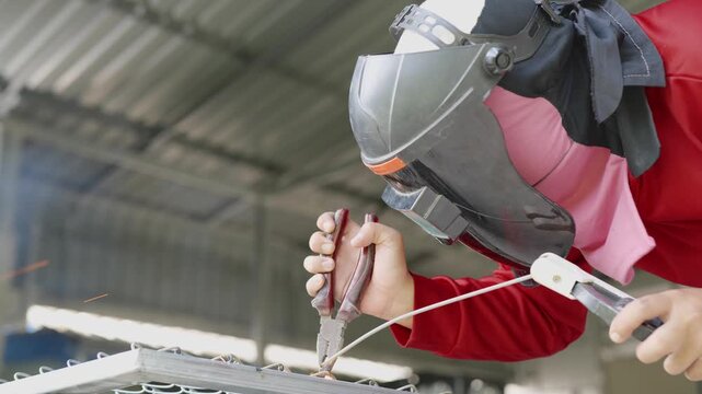 Close-up shot of a worker wearing a protective helmet and red safety sleeves, using a tool to weld or grind a metal wire mesh grille in an industrial workshop. 