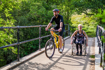 Naklejka premium Man cycling and woman in wheelchair enjoying a sunny day on a bridge.