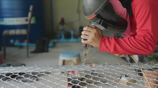 Close-up shot of a worker wearing a protective helmet and red safety sleeves, using a tool to weld or grind a metal wire mesh grille in an industrial workshop. 
