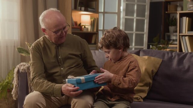 Medium long shot of Caucasian senior man giving gift box to grandson sitting on sofa, boy opening present and revealing toy solar system model. Footage ideal for family bonding and educational ads