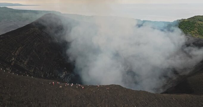Tourists people standing on the dark ash rim of Mount Yasur volcano, observing the active crater while thick white smoke rises in the sky. Travel and explore Vanuatu. Aerial view drone flight panorama