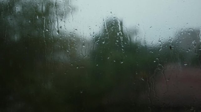 Raindrops stream down a window pane during a heavy downpour, creating a blurry and distorted view of the green trees outside. This moody shot captures the cozy, melancholy atmosphere of a stormy day.