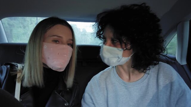 Two women wearing face masks travel sitting in driving car backseat during travel