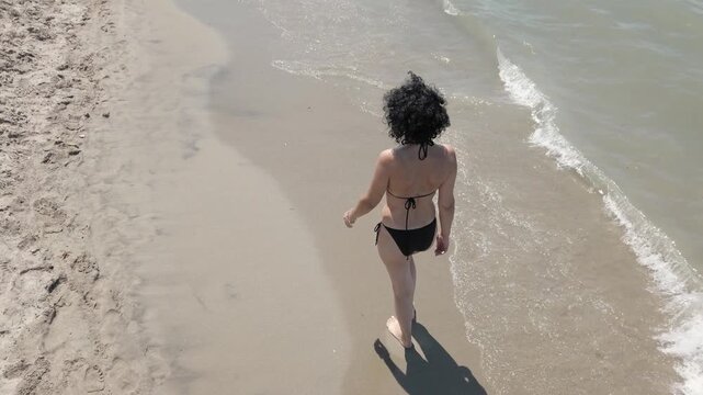 Aerial view of sexy woman in bikini walking along a sandy beach by the water.