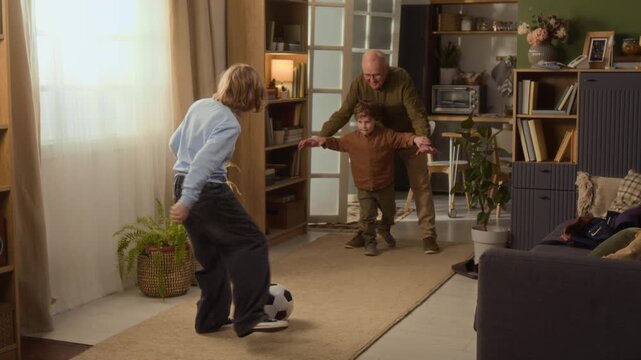 Long shot of senior Caucasian grandfather playing soccer with two grandsons in living room, kicking ball and celebrating. Video ideal for retirement planning or family-oriented marketing campaigns