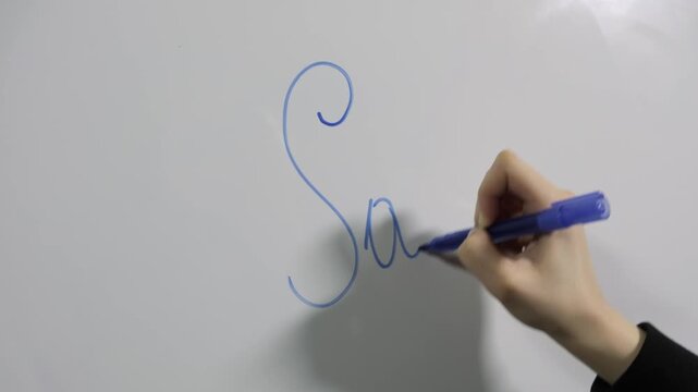 Close-up of a girl's hand writing the word 'sales' on a white board with a blue marker
