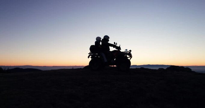 Two individuals riding an ATV at sunset with highlighted silhouettes against colorful sky at twilight