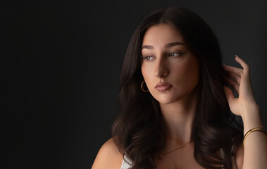 Fototapeta premium Brunette woman looking aside, touching hair, wearing makeup and jewelry on dark background