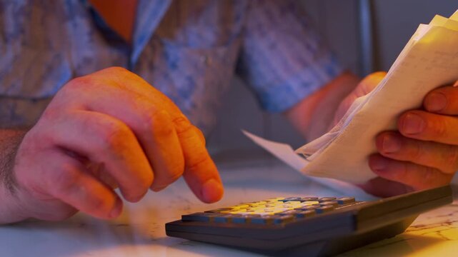 Low angle close-up of man studying family budget expenses by flipping through receipts. Desk lamp light, shallow DoF. Metaphor for home accounting, tracking household costs and financial crisis.