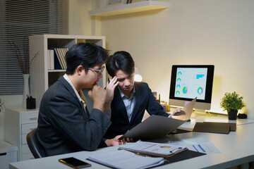 Fototapeta premium Two Asian Business Professionals Reviewing Documents and Data Charts Late at Night in Modern Office
