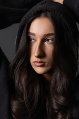 Fototapeta premium Young brunette woman looking away while posing in black hoodie on gray background