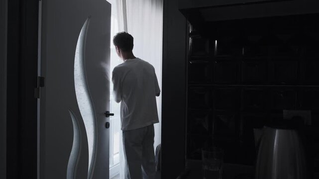 Young white tenant checking curtain, dark interior doorway, cautious hand on fabric, scanning outside for intruder, tense silhouette by decorative divider,