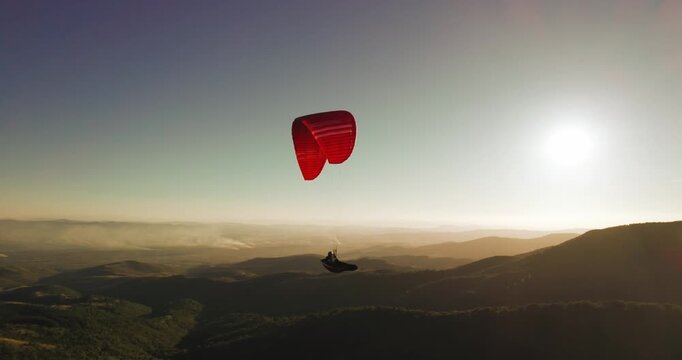 Men with red paraglider soaring gracefully over scenic mountain range and valley at sunset
