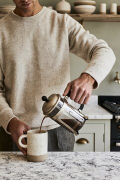 Young man in cashmere sweater pours coffee from a French press into a stoneware mug in a modern kitchen with sage green walls
