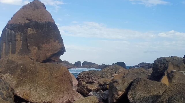 Scenic view of massive rock formations and crashing waves on a tropical rocky beach under blue sky.