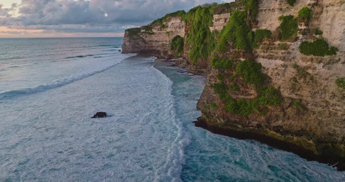 Rugged limestone cliffs covered with green foliage meet the powerful ocean waves crashing along the beautiful coastline of Bali, Indonesia, under a dusky sky at sunset. Aerial view drone panorama