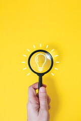 Hand holding magnifying glass over light bulb icon on yellow background, insight and creative idea search concept © Katrin_Primak