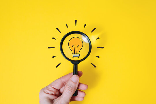 Hand holding magnifying glass over light bulb icon on yellow background, insight and creative idea search concept