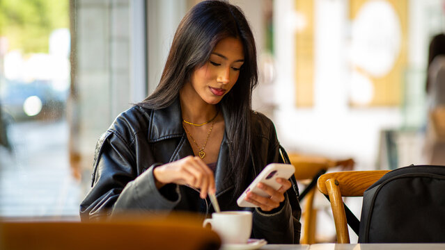 Woman using smartphone while stirring coffee in cafe