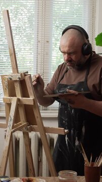 Portrait of a bearded Caucasian millennial man wearing headphones, standing near an easel, holding a palette and painting. Concept: Hobbies and leisure