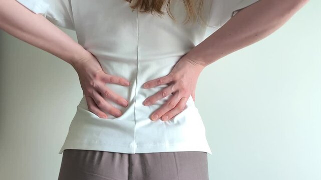 Woman grips her lower back with both hands suffering from sharp pain. She holds her spine tightly while bending slightly forward in discomfort. Concept of severe backache, muscle spasm and