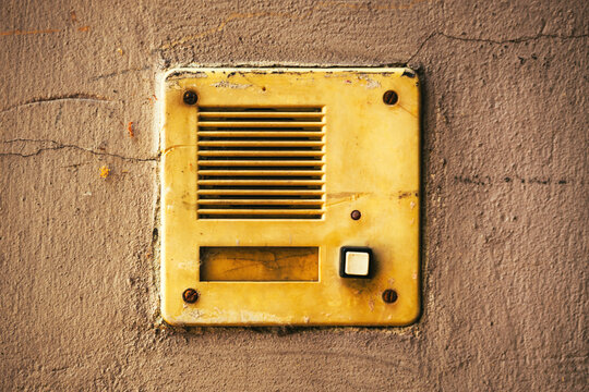 Weathered vintage yellow intercom doorbell speaker with push button mounted on a cracked concrete wall, showing rust, peeling paint and textured surface
