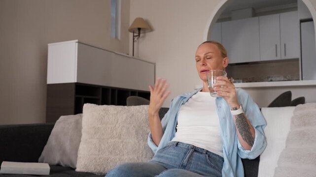 Woman sit in living room feeling overheated during summer heat, fanning herself with hand and drinking water from glass to cool down at home.Hydration, heat discomfort, home cooling struggle concept