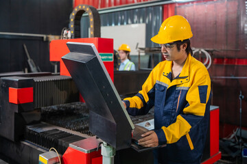 Fototapeta premium Worker in a yellow and blue work jacket operates a large industrial machine. They wear a yellow hard hat and use a control panel. Another worker in a yellow hard hat is visible in the background