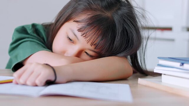 Close up selective focus of a tired Asian kid girl resting her head on the desk. Stressed and exhausted from studying too much homework in bright daylight. Education burnout and overwork concept.