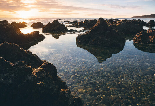 A rocky coastline at sunrise with dark volcanic stones, clear tide pools reflecting the sky, and calm ocean water under a soft morning light