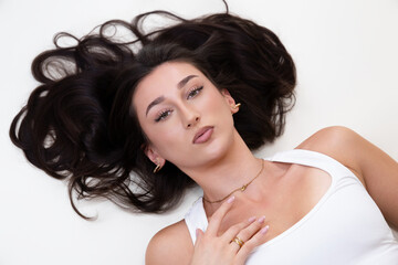 Naklejka premium Beauty and fashion portrait of a young woman with long dark hair, natural makeup, and elegant jewelry. The female model is lying down, looking directly at the camera, her features and accessories. Thi
