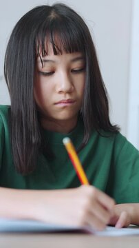 Vertical video close up and selective focus showing Asian kid girl concentrating as she writes in her notebook with a pencil. Bright daylight fills the room while she is doing her homework.
