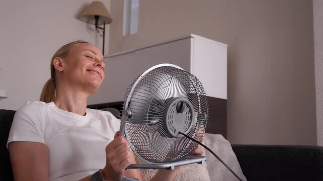 Woman suffering from extreme heat at home during abnormal heat wave, sitting on sofa and using electric fan to cool down in dangerously hot indoor temperature.Extreme heat, climate change heatwave