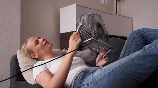Woman lying on sofa in home during extreme heat wave, cooling her face with electric fan and feeling relief from dangerously hot indoor temperature.extreme heat,  climate heatwave concept