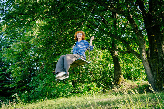 Happy woman in a hat and sunglasses swinging on a swing in a sunny summer forest