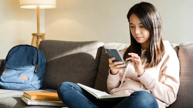 Settling student in light-pink hoodie tapping smartphone and checking notebook on sofa to study