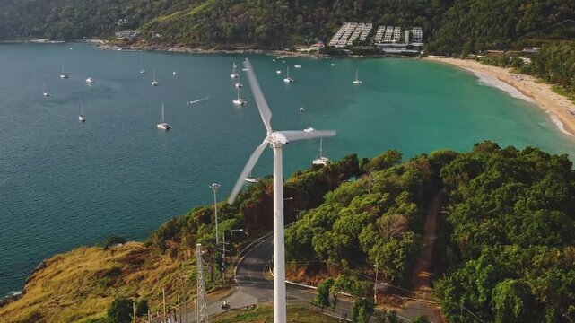 Wind turbine spinning on a green hill overlooking turquoise Andaman Sea while numerous sailboats and yachts anchor in the bay and tourists relax on the sandy beach in Thailand. Aerial view drone shot
