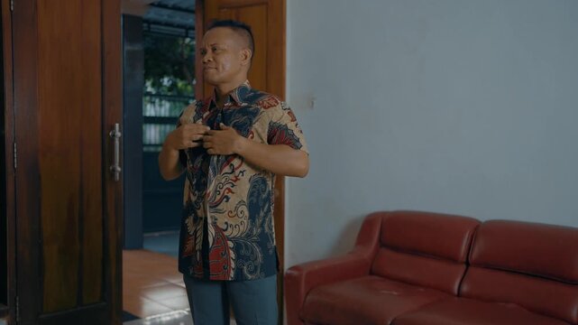 Adult Asian man buttoning batik clothes shirt in fitting room at home.
