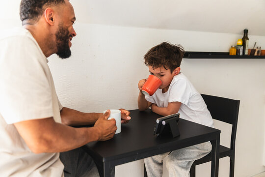 Boy drinking from mug with father watching phone