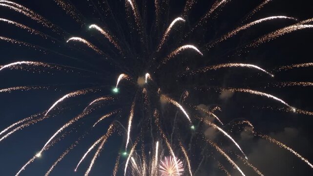 Firing shells detonating into growing bursts in night sky, emitting trails, smoke, building finale