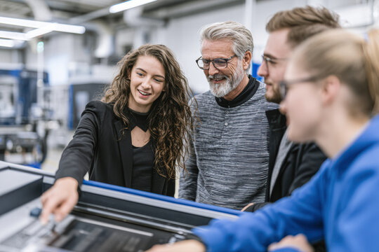 A group of four diverse professionals collaborate around a large printer or copier in a modern office setting, engaging enthusiastically with the machine's controls.