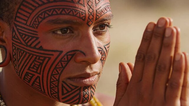 Turning, tattooed man holding pressed palms in ritual pose on sunny sand, wearing plug and beads