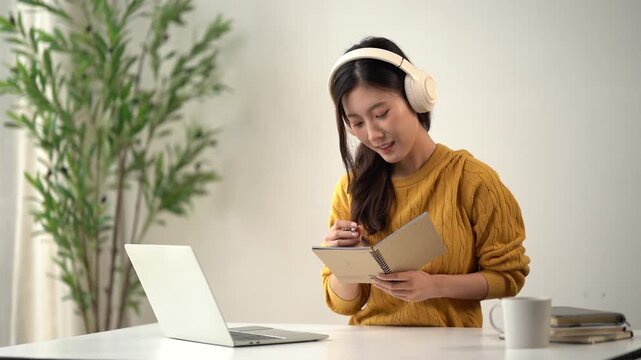 Concentration and Learning: A young woman with headphones on is engrossed in a book while a laptop is open on the desk, indicating focused learning or remote study.